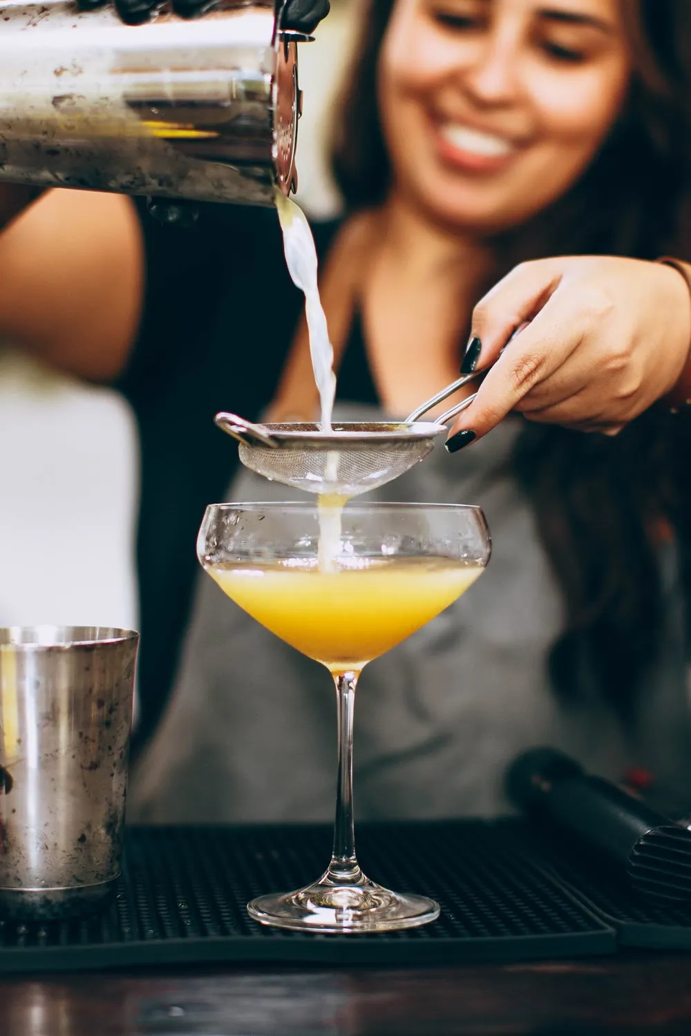 Bartender sorrindo enquanto coa um drink amarelo numa taça